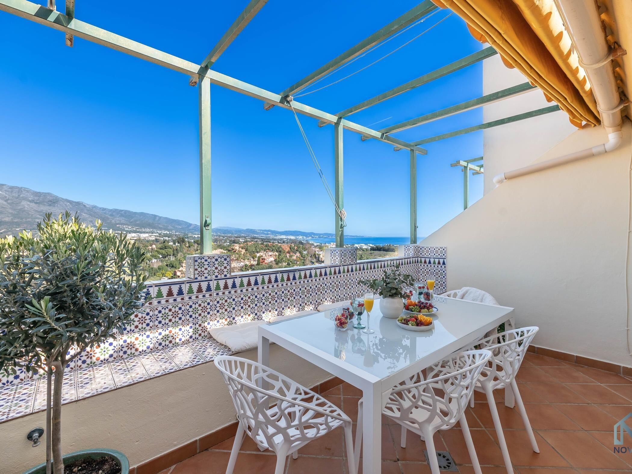 A terrace with a table set for two, white chairs, a view of the sea, and a tiled wall with a geometric pattern.
