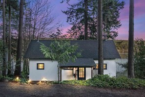 Una casa blanca con techo oscuro, rodeada de árboles altos y una masa de agua.
