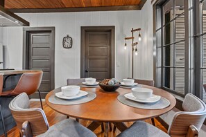 Un comedor con una mesa redonda de madera dispuesta para cuatro personas, rodeada de sillas. Se ven dos puertas al fondo, una con un reloj encima.