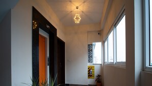 A hallway with a checkered floor, a potted plant, a fire extinguisher, and a window with a view of buildings. - Sarvam Serviced Apartment (Coimbatore)