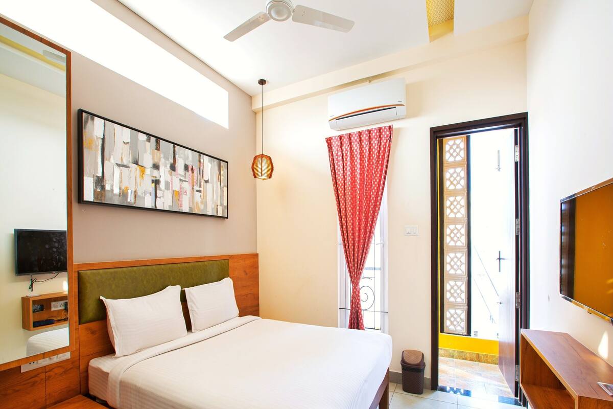 A hotel room with a bed, a television, a ceiling fan, an air conditioning unit, and a window with a red curtain.