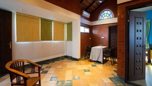 A spacious room with a high ceiling, a wooden bench, a table with chairs, and a large window with blinds. - Sarvam Serviced Apartment (Coimbatore)