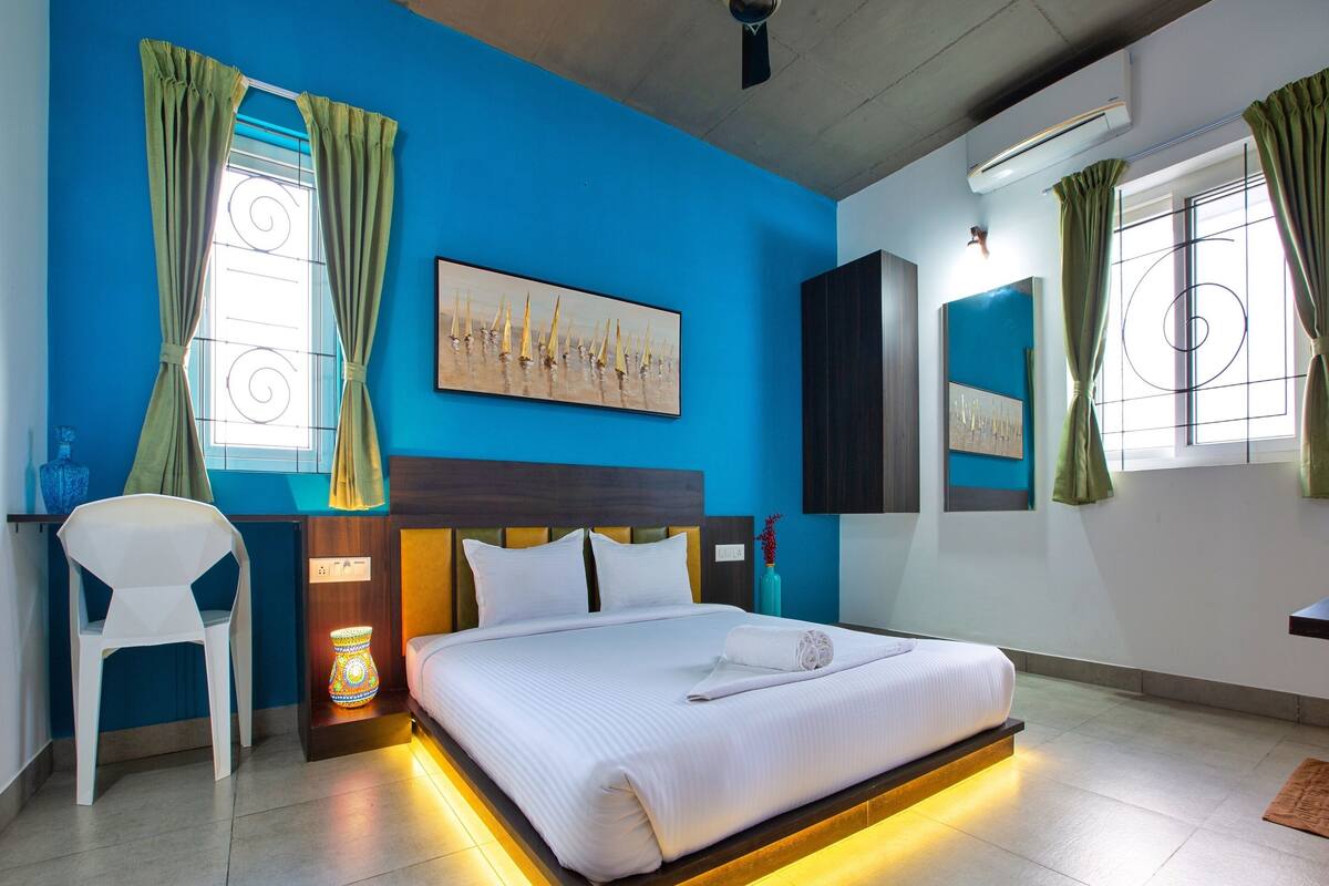 A modern hotel room with a large bed, a chair, a painting, and a ceiling fan.