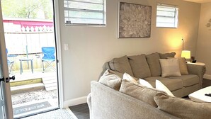 A living room with a beige sofa, a coffee table, and a lamp. There is a door leading to an outdoor area with a patio and a blue chair.
