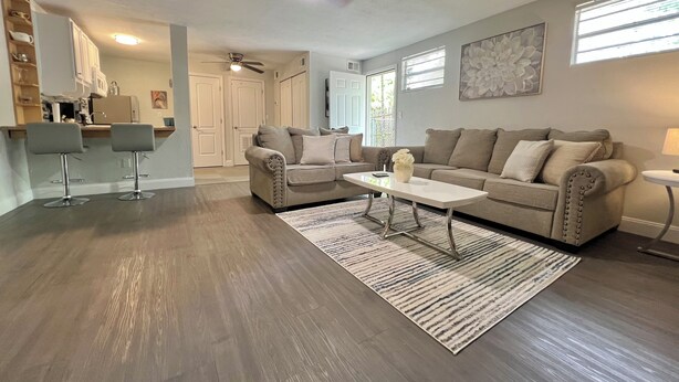 A living room with a sofa, coffee table, and two bar stools. There is a ceiling fan, a rug, and a window with blinds.