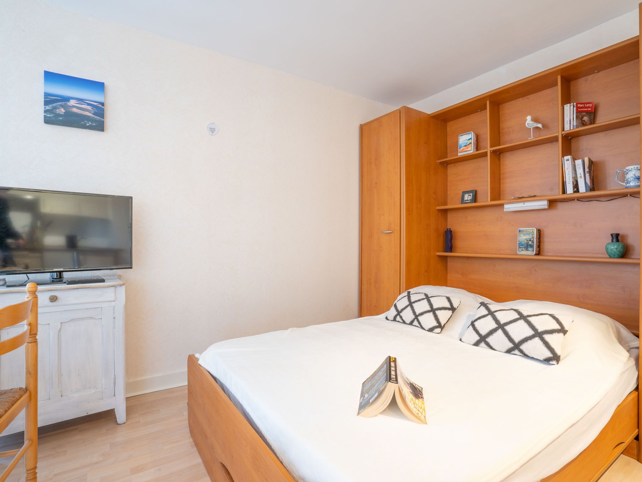 Een slaapkamer met een bed, een televisie op een dressoir, een boekenkast en een schilderij aan de muur.