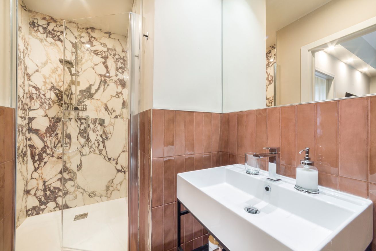 A bathroom with a marble shower wall, a white sink with chrome fixtures, and a mirror above the sink.