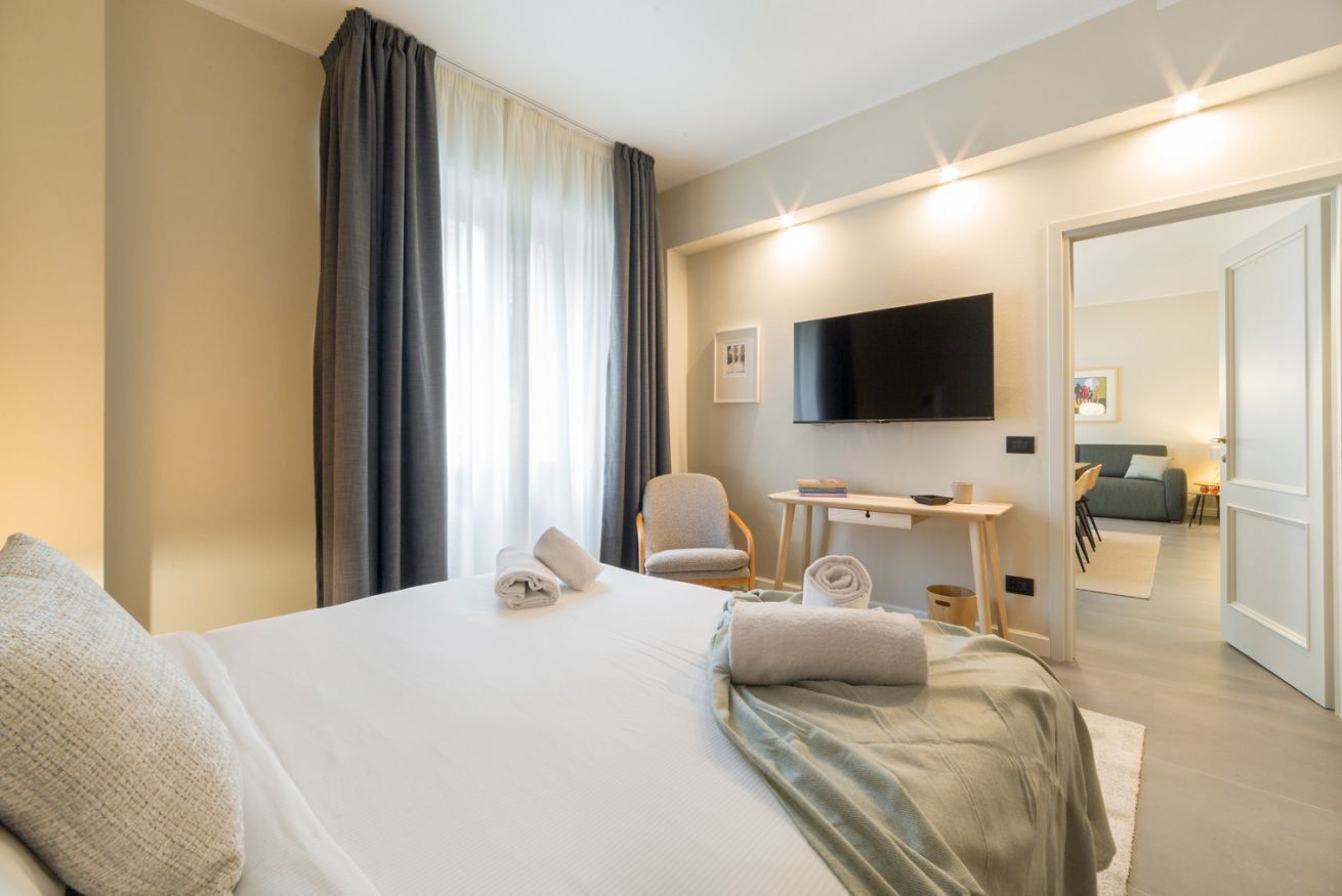 A hotel room with a bed, a desk, a chair, a TV, and a view of another room.