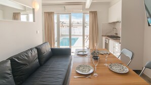 Un comedor con una mesa de madera dispuesta para dos personas, un sofá de cuero negro y vistas a una piscina y un balcón a través de grandes ventanales.