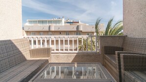 Un balcón con mobiliario de mimbre, una mesa de vidrio y vista a un edificio con balcones y palmeras.