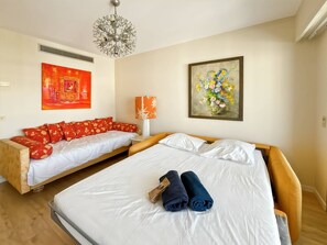 A hotel room with two beds, a chandelier, a painting, and a red sofa. - Three Bedroom Sea View-ORS7 (CANNES)
