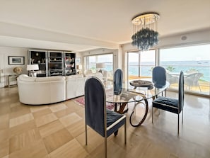 A modern living room with a glass dining table, white sofas, and a view of the sea.