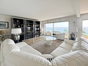 A modern living room with a glass coffee table, white leather sofa, and a view of the ocean through large windows.