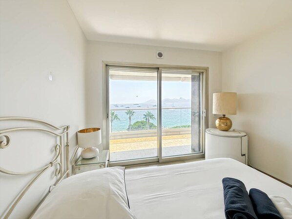 A hotel room with a bed, a bedside table with a lamp, a window with a view of the sea, and a balcony. - Three Bedroom Sea View-ORS7 (CANNES)