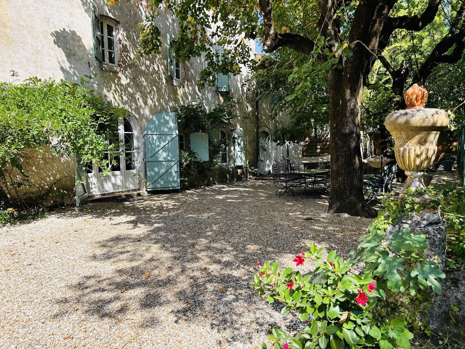 Un patio con un sendero de grava, un edificio rústico con una puerta azul y un árbol grande.