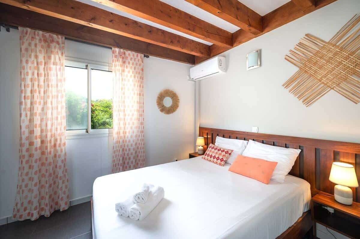Ein ordentlich bezogenes Bett mit weißen Leinen und orangefarbenen Kissen, ein Holz-Kopfteil, eine Wand-Klimaanlage und ein Fenster mit gemusterten Vorhängen.