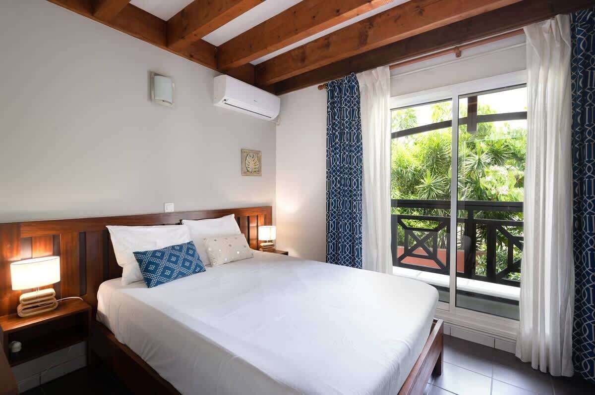 A hotel room with a wooden headboard, a large bed with white bedding, two bedside lamps, an air conditioning unit, and a balcony with a view of greenery.
