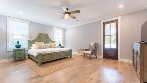 A bedroom with a wooden floor, a bed with a headboard, a chair, a nightstand, a lamp, a ceiling fan, and a door.