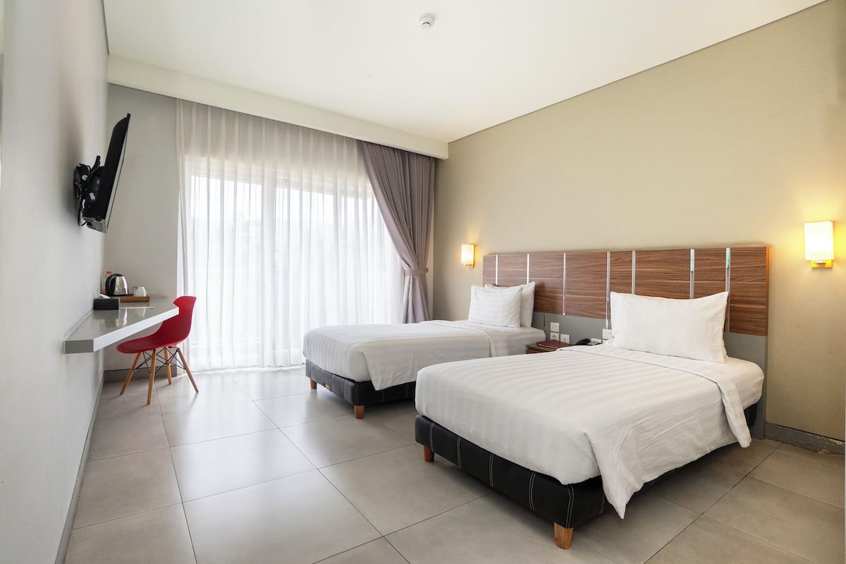 Habitación de hotel con dos camas, un escritorio con silla, televisor montado en la pared y ventana con cortinas.