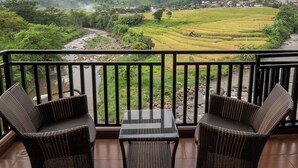 A balcony with wicker chairs and a table, overlooking a river and green fields with a village in the distance.