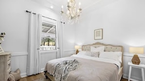 A bedroom with a large bed, a chandelier, a window with curtains, a bedside table with a lamp, and a dresser. - The Dawning Farmhouse (Jaspers Brush)