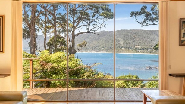 A room with a large sliding glass door leading to a deck overlooking a coastal landscape with trees and hills. - Osprey Lodge - Bungalow & House (Eaglehawk Neck)