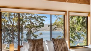 A bedroom with a large window offering a view of a lake and trees. There is a bed with white linens and two patterned pillows. A small table with a lamp is visible to the left of the bed. - Osprey Lodge - Bungalow & House (Eaglehawk Neck)