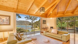 A spacious living room with a wooden ceiling, large windows offering a scenic view of the sea and hills, and a cozy seating area with a sofa and a coffee table. - Osprey Lodge - Bungalow & House (Eaglehawk Neck)