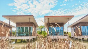 Twee moderne huizen met grote ramen en balkons, gelegen boven water met hoog gras op de voorgrond.