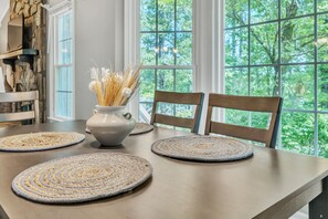 Una mesa de comedor con individuales tejidos, un florero con plantas secas y vistas a árboles a través de grandes ventanales.