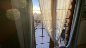 Quarto com janela coberta por cortinas finas, piso de azulejos e vista para área externa com plantas em vasos e uma cadeira.