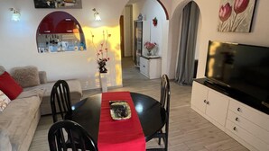 Uma sala de estar moderna com uma mesa de jantar, cadeiras pretas, um sofá, uma televisão e uma cozinha ao fundo.