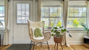 Et værelse med trægulv, en fletstol med pude med bladmotiv, en potteplante og udsigt til et hus gennem store vinduer.
