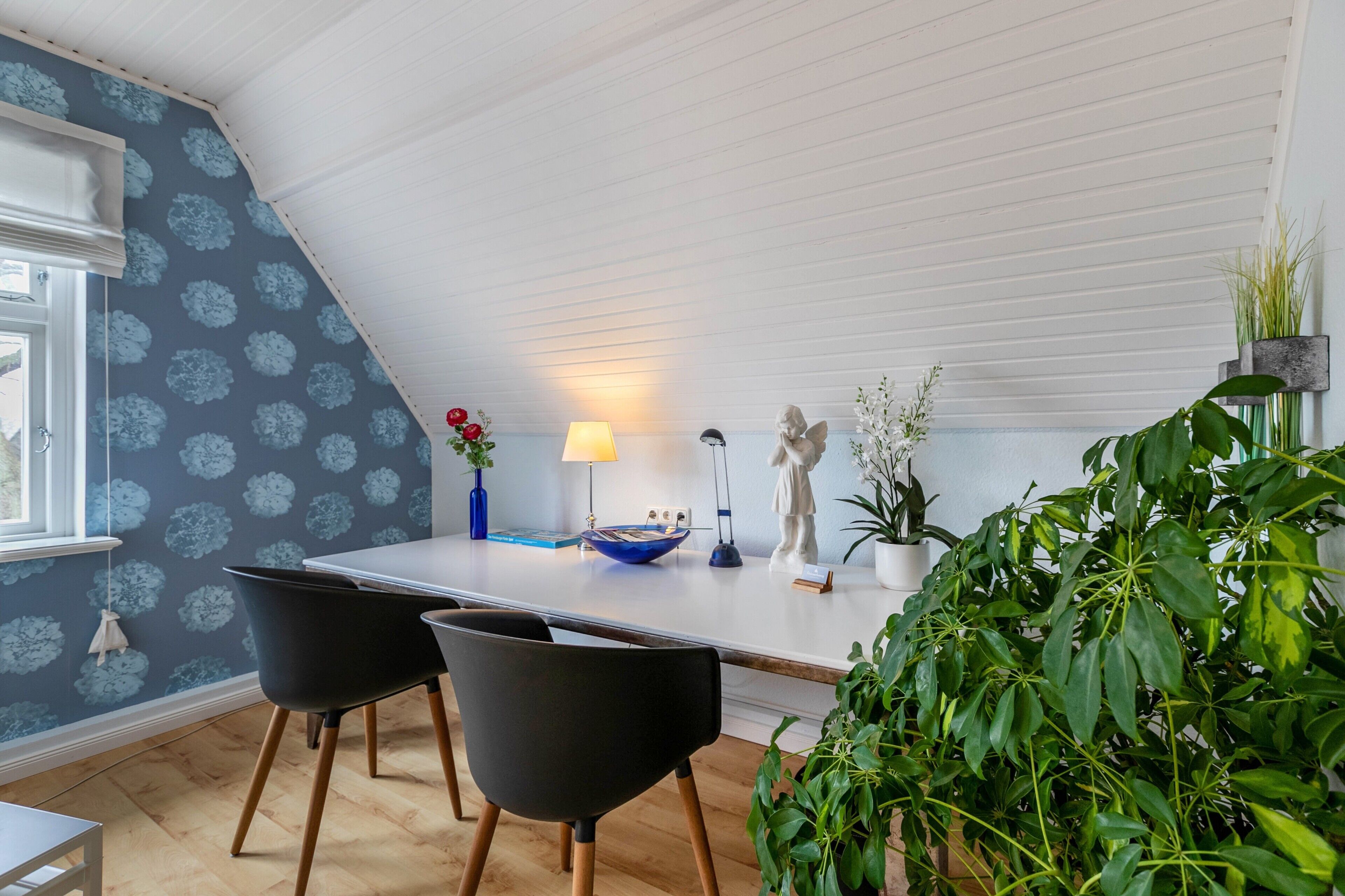 A dining area with a white ceiling, a blue floral wallpaper, a table with chairs, a lamp, and decorative items.