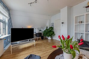 Uma sala de estar com televisão de tela plana, piso de madeira, uma planta em vaso, um vaso com tulipas vermelhas e uma porta branca.