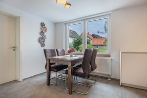 Ein Essbereich mit Tisch, Stühlen, einer Blumenvase, einem Fenster mit Blick auf Häuser und einer Heizung.