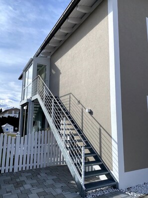 A modern building with a metal staircase leading to an upper level, a white picket fence, and a paved ground.