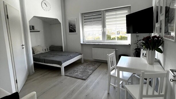 A compact room with a bed, a desk, a chair, a TV, and a window with blinds. - Studio Apartment 'Lorenz' with Wi-Fi (Olching)