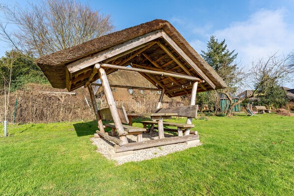 A rustic wooden structure with a thatched roof, surrounded by a grassy area and trees.