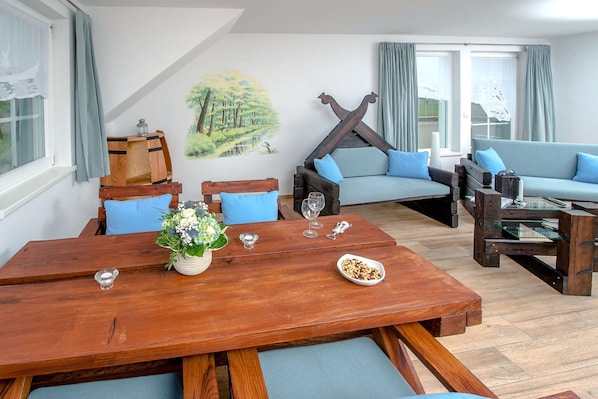 Un salon avec une table à manger en bois, des canapés bleus et un tableau représentant une forêt.