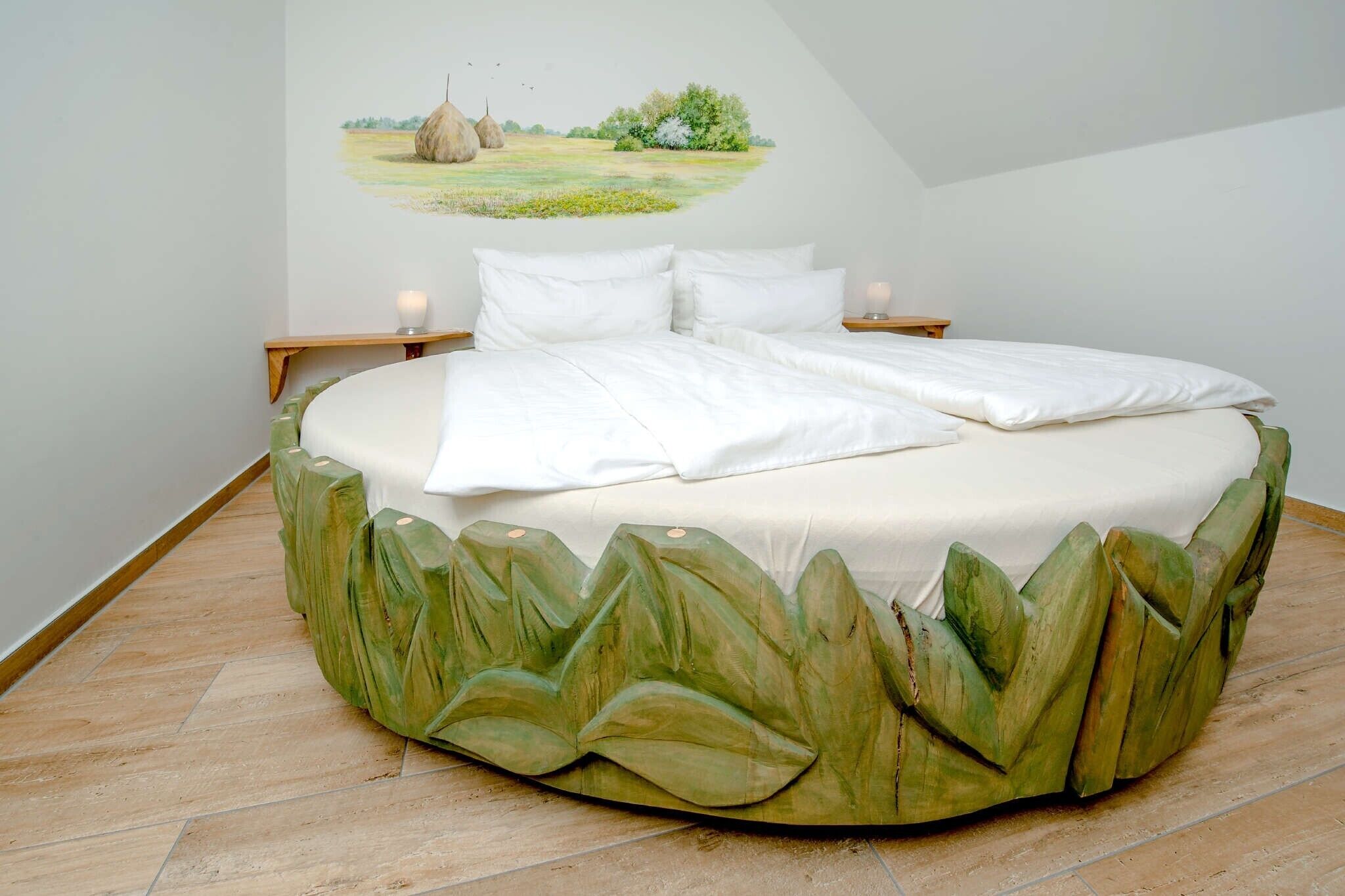 Un lit rond doté d’une base ornée de motifs de feuilles, d’une literie blanche et d’une fresque murale représentant un paysage.