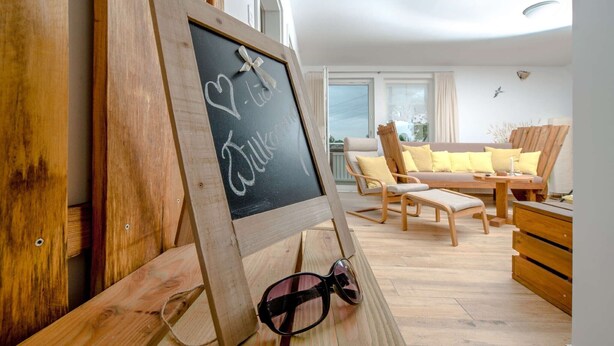 Una pizarra de madera con un corazón y texto, un par de gafas de sol, una mesa de madera y una zona de estar acogedora con cojines.