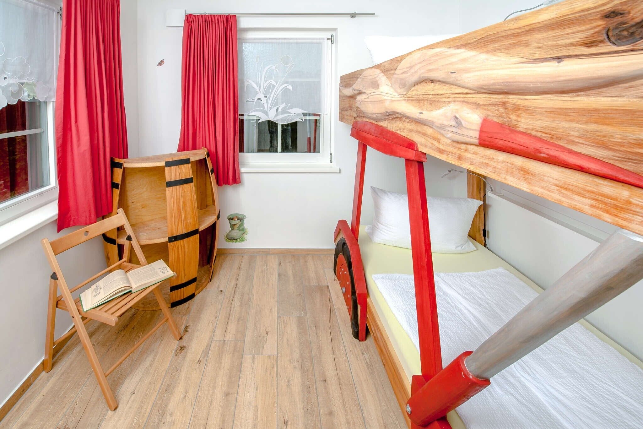 Une pièce avec un lit superposé, une chaise en bois, un tonneau et une fenêtre ornée de rideaux rouges.