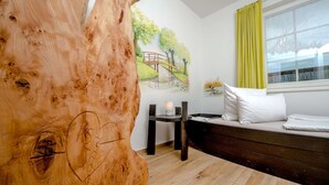 Ein Zimmer mit einer großen Holzskulptur, einem Bett mit weißer Bettwäsche, einem kleinen Tisch mit einer Kerze und einem Wandbild einer Brücke.