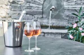 Ein Edelstahlbehälter mit einem Löffel, zwei Gläser mit einem rosa Getränk und eine Mauer aus Stein im Hintergrund.