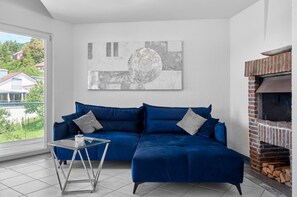 Ein Wohnzimmer mit einem blauen Sofa, einem Glastisch, einem Kamin und Blick auf Häuser durch ein großes Fenster.