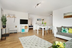 A modern living room with a flat-screen TV, a dining area with a white table and chairs, a grey sofa with cushions, and a large potted plant.