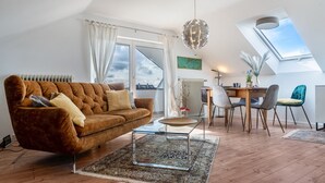 Un salón estilo loft con un sofá marrón, una mesa de centro de vidrio, una zona de comedor con una mesa y sillas de madera, y un tragaluz.