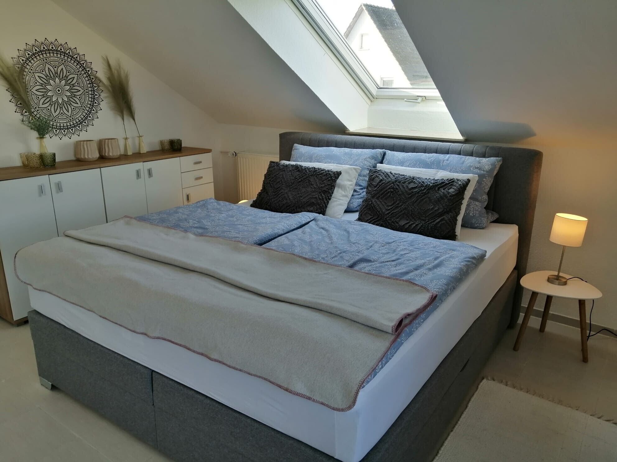 Quarto com claraboia, cama com várias almofadas, mesa de cabeceira com abajur e um objeto decorativo na parede.