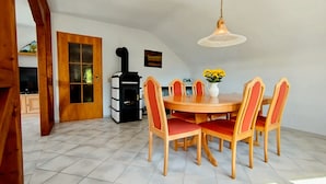 Un comedor con una mesa redonda de madera, seis sillas, un jarrón con flores amarillas y un estufa de leña negra.